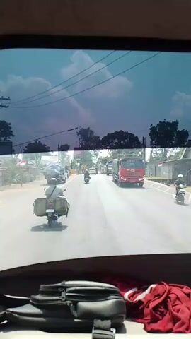 A video displays a view through a vehicle's rear window, capturing a road with traffic. In the foreground, a dark bag and some red fabric are visible on the vehicle's interior. The road is paved and appears to be a two-lane highway with vehicles traveling in the same direction. A motorcycle with luggage is in the left lane, with other motorcycles and cars further ahead. Several trucks are also present, including a red truck in the right lane and a white truck further down the road. Trees and buildings line the sides of the road. The sky is cloudy with patches of blue. The footage appears to be taken from a moving vehicle.