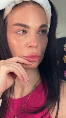 This is a close-up, vertical video of a young woman with long, dark hair styled in pigtails and wearing a white headband. She is wearing a bright pink ribbed tank top and a gold chain necklace with a green and red pendant. Her hand is resting on her chin, with her fingers gently touching her lips. She has full, glossy lips, and her makeup appears to be natural, with a focus on glowing skin and defined eyebrows. Her eyes are a striking green. The lighting suggests an indoor setting, possibly a bathroom or dressing area, as evidenced by the soft light and a portion of what appears to be a black surface in the background, possibly a cabinet or door. There are no discernible weather cues or time of day indicators, though the lighting is not harsh, suggesting it's not direct midday sun. The overall mood conveyed is one of quiet contemplation or a moment of self-reflection, perhaps before or after a beauty routine. There is no visible text in the media.