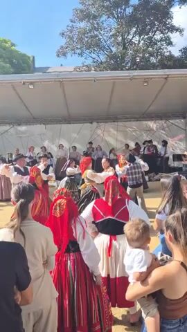 A group of people in traditional folk costumes are dancing on a stage outdoors under a blue sky with some trees visible. The dancers on stage are mostly women wearing red and white dresses with colorful striped skirts, and men in white shirts and vests. They are performing a lively dance, with some pairs holding hands. In the foreground, facing away from the camera, are several spectators, including a woman holding a young child. The spectators are dressed in casual modern clothing. The stage is covered by a white canopy.