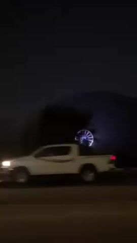 The video shows a Ferris wheel lit with blue lights at night. It is positioned against a dark hill or mountain. The video is shot from a moving car. A car passes in the foreground.