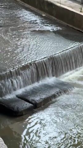 This is a video clip showing a section of a water channel with a small waterfall. The water is flowing, creating ripples and a cascade over a concrete ledge. In the foreground, two dark, rectangular objects, likely concrete blocks or slabs, are submerged in the water just below the waterfall. The water appears somewhat murky. The surrounding environment includes concrete walls on the sides and top of the channel, with some greenery visible at the very top edge of the frame. The lighting suggests it is daytime, possibly overcast, as there are no harsh shadows. The overall scene is industrial or urban infrastructure, likely part of a drainage or water management system. No people or animals are visible. There is no discernible text within the video. The Ampang Jaya Municipal Council, Malaysia, is mentioned as a location context, suggesting this might be a part of their infrastructure.