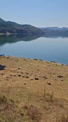 A vertical video displays a wide shot of a calm lake with hills in the background under a clear blue sky. The foreground is a dry, rocky, and grassy embankment. In the middle ground, the water is a deep blue, reflecting the sky and distant hills. Several dark specks, possibly birds, are visible on the water. A small white boat is moored on the right side of the lake. The camera pans slightly to the right.