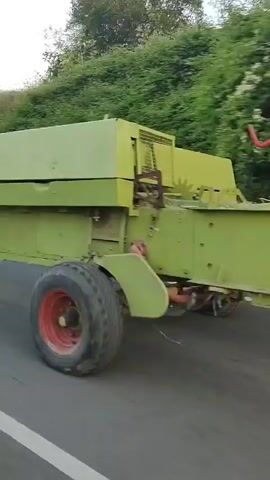 The video captures a side view of a green trailer being pulled by a red tractor on a paved road. The trailer is large and boxy with a visible tire on the right side. The tractor is a classic model with large rear wheels and smaller front wheels, and it has a yellow roll cage. The tractor's headlights are on. The road is lined with trees and vegetation on a slight incline to the left of the frame. The scene appears to be outdoors in daylight.