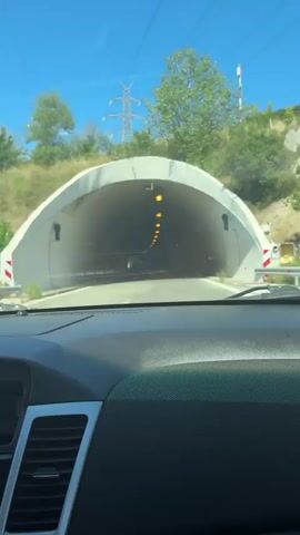 The video is filmed from inside a vehicle, looking through the windshield at the entrance of a tunnel. The tunnel is illuminated by a series of yellow lights along its ceiling. The vehicle is approaching the tunnel, and the exterior environment, which appears to be a sunny day with some trees and sky visible, is quickly obscured as the vehicle enters the tunnel. The interior of the tunnel is a dull, concrete gray, and the lighting becomes more prominent as the vehicle progresses deeper. The video continues for a few seconds as the vehicle moves further into the tunnel, with the distant opening of the tunnel becoming brighter.