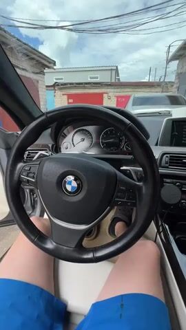 This media file shows a first-person perspective from inside a BMW. The subject is seated in the driver's seat, wearing blue shorts and light-colored shoes. The steering wheel, dashboard with a speedometer and tachometer, and center console are clearly visible. The car appears to be parked in an outdoor setting, with buildings and power lines visible through the windshield. The weather seems overcast, with a cloudy sky. The scene suggests a mundane moment, possibly before or after a drive, with no strong emotions conveyed. The visible text includes "BMW" on the steering wheel and "AIRBAG." There are also numbers "133" and "134" visible on the buildings in the background. The location context is Veteran-2, Russia. The activity is simply being inside the car, with the engine starting up and revving at the end of the clip.