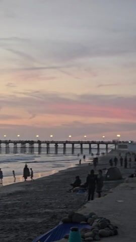 The video shows a beach scene at sunset. A pier stretches into the ocean, illuminated with lights. People are visible along the beach, some walking, some standing in the water, and some sitting on the sand. The sky is colored with pink and orange hues. Waves are lapping onto the shore, reflecting the colors of the sky. There are some rocks and what appears to be a blue tarp in the foreground.
