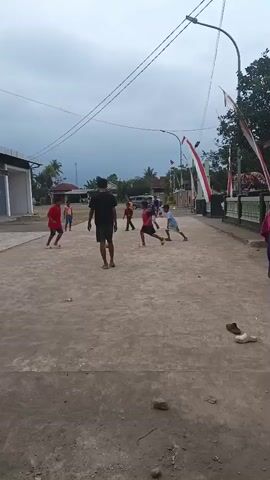 The video shows a group of children playing soccer in a street. There are at least five children visible, dressed in casual clothing, with one boy wearing a red shirt and black shorts, another in a red shirt and blue shorts, and a third in a red shirt and dark shorts. One of the children is kicking a yellow soccer ball. The street appears to be paved with concrete and dirt, and there are houses and trees on either side. Red and white flags are displayed on poles along the side of the street. The sky is overcast.