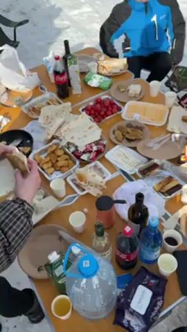 The video shows a festive outdoor gathering on a snowy landscape. A folding table is centrally positioned, laden with a variety of food and beverages. Dishes such as sliced cheese, cherry tomatoes, pastries, and stacks of lavash bread are arranged on the table. Drink options include Coca-Cola, wine, and water, all neatly placed around the various food items. 

Two people are visible. One is seated, wearing a blue jacket and dark pants. The second person stands next to the table and appears to be preparing food, assembling ingredients in the lavash bread. Other chairs are visible around the table. The ground is completely covered with snow. The overall impression is that of a lively, informal gathering outdoors, with people enjoying food and drinks in a snowy setting.