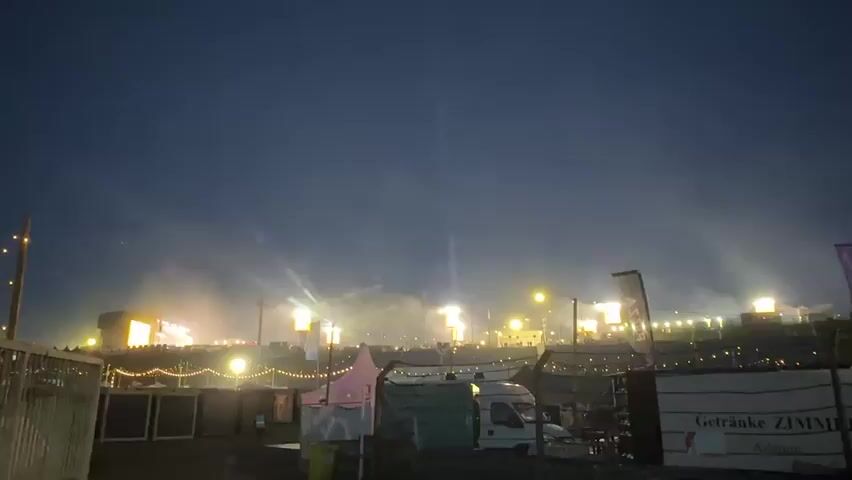 The video shows an outdoor night scene at what appears to be a festival or concert. The sky is dark blue. In the foreground, several temporary structures, possibly stalls or tents, are visible, illuminated by string lights. A white van is parked. On the far right, a dark banner with the text "Getränke ZIMMER" and "Adenau" is displayed. In the midground and background, a large stage or multiple stages are active with a dynamic light show featuring pink, red, white, and yellow lights. Beams of light project into the sky, and smoke or fog rises from the stage area. Silhouettes of people are discernible in the distance near the stages.