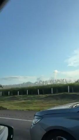The video depicts a silver sedan driving on a road, as viewed from another car. The camera is positioned inside the second car, looking out the side window. The sedan moves at a moderate speed, with a clear view of its side profile and rear. Inside the sedan, at least one person can be seen in the driver's seat. The background outside the car features a rural landscape with green fields, trees, and a mountain range in the distance under a clear blue sky with scattered clouds. A white picket fence lines the roadside. The lighting suggests it is daytime.