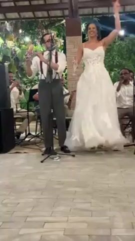 A wedding band, including a flute player, trumpeter, guitarist, singer, and other musicians, is playing music for a wedding celebration. A bride in a white, floral lace gown raises her arm and dances with a champagne glass in her hand. A wedding guest in a long, light-green dress dances in front of the band, showing the dress’s back and strap detail. The musicians are wearing white shirts and most are seated. A black speaker is beside the guitar and bass. The venue has a stone tile floor, a partially covered, open ceiling, and is decorated with string lights and greenery.