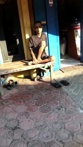 The video shows a street scene in Tenggulunan, Indonesia. The foreground features a patterned red and grey tiled walkway. In the mid-ground, a man is seated on a low stool outside a building with a bright blue door. He is wearing a dark t-shirt and shorts. Next to him is a bicycle wheel. Behind the man, the building appears to be a shop or workshop, with a display counter filled with various items, including bottles and cleaning supplies. A large water cooler is visible. The shop also has shelves with products and a cash register. Sunlight casts strong shadows on the ground, suggesting it is daytime, likely late morning or early afternoon given the angle of the sun. The weather appears to be sunny and warm. The overall atmosphere seems relaxed, with the man casually observing his surroundings.