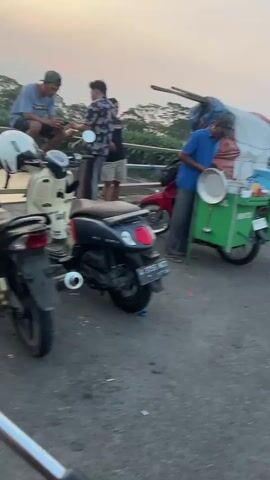 The video shows a street scene at dusk. Several people are present, some of whom are interacting with each other. Motorcycles are parked in the foreground. In the background, a vendor is operating a food cart. The environment appears to be outdoors, possibly near a body of water given the presence of what looks like a bridge or elevated structure in the background. The lighting suggests it is either sunrise or sunset.