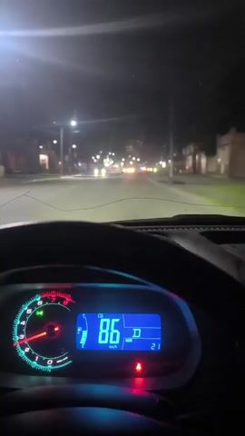 This is a first-person perspective video of driving at night on a paved road. The dashboard of a car is visible in the foreground, displaying a speedometer that reads 87 km/h, then 89 km/h, and finally 92 km/h. The road ahead is illuminated by streetlights and the headlights of other vehicles, with buildings and trees lining the sides of the road. The sky is dark, indicating nighttime.