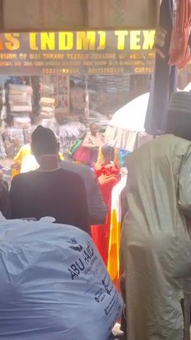 The video shows the exterior of a textile shop, "AINAS (NDMJ) TEXTILES," located in the Mai Turare Textile Dealers of Veils, Kwari Market, New Shop O.P.Q/Madawa. There are people browsing and walking around the shop. The shop has a variety of textiles, including dresses on hangers, stacks of colorful fabric, and bags. The phone numbers 08085182334 and 08173000440 are visible on the storefront sign. The environment appears busy and crowded, indicative of a market setting.