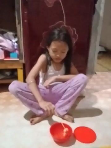 A young girl sits cross-legged on the floor, wearing a white top and lavender pants with white designs. A red plastic cup sits on the floor next to her feet, its lid next to it. The girl appears to be manipulating something small with her hands. To her left is a wooden cabinet with a dark red door, and a partially visible shelf with items in it. The floor is a light-colored tile. The camera angle is overhead, looking down at the girl and her surroundings.