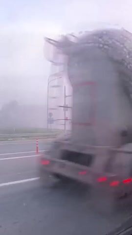 The video shows a foggy intersection with a traffic light. The traffic light is red, and a white van is seen crossing the intersection. There are also several orange traffic cones on the road. The sky is overcast with pink and purple hues, suggesting it is either sunrise or sunset.