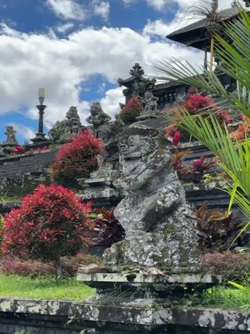 The video shows a temple in Bali, Indonesia. The temple is built on a hillside, and there are many stone statues and carvings. The statues are covered in moss and lichen, and the plants are green and red. The sky is blue with white clouds.