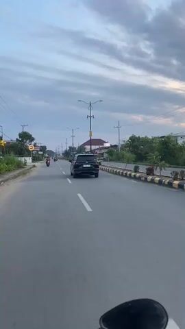 The video shows a view from a vehicle, likely a motorcycle, moving along a paved road. The road has clear white lane markings and a yellow and black striped median barrier on the right side. There are streetlights lining the road at intervals. In the distance, a black SUV is driving in the same direction. On the left side of the road, there are some buildings, trees, and a few motorcycles with riders. The sky is cloudy, with a mix of grey and lighter clouds, suggesting it might be dawn or dusk. The overall environment appears to be a suburban or semi-urban area.