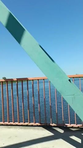 This media file shows a view from a bridge looking out over a body of water. The scene is set on a clear, sunny day, indicated by the bright blue sky and the strong shadows cast by the bridge's structure. The water appears calm and blue, with gentle ripples on its surface. A reddish-orange railing runs along the edge of the bridge, with vertical bars spaced evenly. A large, teal-colored structural element of the bridge dominates the left side of the frame, angling upwards into the sky.

The perspective suggests movement, likely from a vehicle crossing the bridge, as evidenced by the slight blur and the changing angles of the bridge's structure in subsequent frames. The overall impression is one of a serene, scenic drive across a bridge, with the focus on the expansive sky and the tranquil water. There are no people visible in the frame. The setting appears to be a natural environment, possibly a river or a lake, with a distant shoreline visible in the background, hinting at land on the other side. The time of day is clearly daytime due to the strong sunlight and clear visibility. The location context of Gryfino, Poland, suggests this could be a bridge over the Oder River. No visible text is present in the media file.