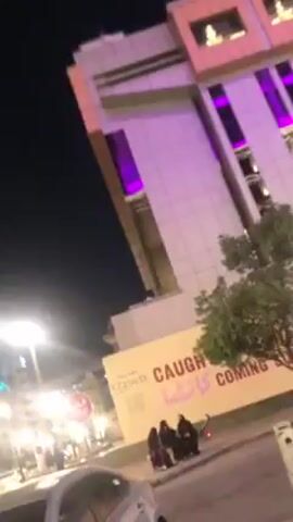 The video shows a nighttime street scene. A light silver-colored car is parked on a brick-paved area, with its left side facing the camera. In the background, a group of people in dark clothing, possibly abayas, stand near a temporary wall with the words "CAUGHT KAITA COMING SOON" written on it. The wall is in front of a tall building that has purple lighting accents. There is a small tree between the wall and the building. A street lamp is visible on the left side of the frame. A yellow painted line is visible next to the parked car.