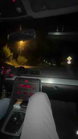 The video shows the interior of a car at night, from the passenger seat's perspective. The dashboard is visible, with red lights illuminating the radio and instrument panel. A hand wearing a light blue long sleeve shirt is seen shifting the gear stick. The driver is wearing gray pants. Streetlights shine through the windshield. The music is playing in the background.