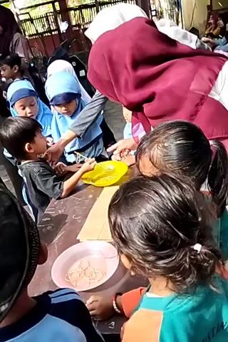A group of young children, some wearing hijabs, are gathered around tables outdoors. One child, wearing a black shirt, is being offered something from a yellow bowl by an adult wearing a burgundy hijab. Another child, wearing a blue hijab, is looking at the interaction. In the background, other children and adults are visible, some holding phones. The ground is paved with interlocking stone tiles. There are motorcycles and a white truck in the background. The scene appears to be an outdoor event or gathering.