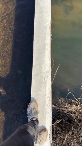 The video shows a first-person perspective of someone walking along a narrow concrete beam. On either side of the beam is water, with visible shadows on the left side.  The person is wearing sneakers and dark pants. There is some dry brush along the right side of the beam.