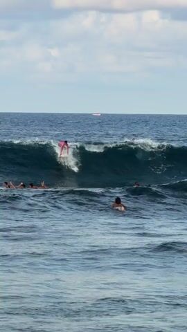 The video shows several people surfing in the ocean under a partly cloudy sky. One surfer, wearing a pink shirt, is riding a moderate-sized wave. Other individuals are either on surfboards or swimming nearby in the water. In the distant background, there is a small boat on the horizon. The water is a mix of dark and light blue hues.