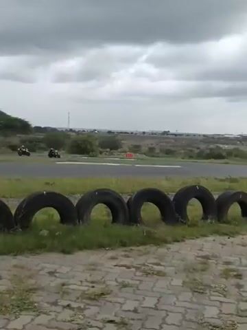 Two motorcyclists race around a paved track. One rider wears a black suit with a red helmet, riding a white motorcycle with black and blue accents. The other wears a black suit and red helmet, riding a motorcycle with a unique multicolored design. The track is lined with grass and tires, and a town is visible in the distance under a cloudy sky.