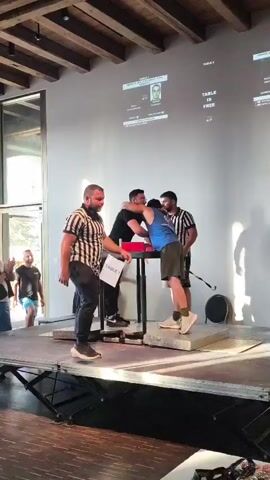 The video shows an arm wrestling match taking place on a stage. Three men are at an arm wrestling table. Two of the men are facing each other, gripping hands over the table, ready to compete. The third man, dressed as a referee in a black and white striped shirt, stands to the left, observing the competitors. Another man in a referee shirt stands behind the competitors on the right. A large screen is projected onto the wall behind the competitors, displaying their names and images. The environment appears to be an indoor venue, possibly a hall or event space, with wooden beams visible on the ceiling. Other people are visible in the background, some standing and watching. The stage is elevated, with metal platforms for the competitors to stand on.