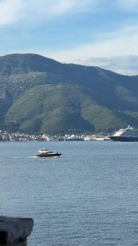 The video shows a small motorboat moving across the water from left to right in front of a docked cruise ship. The mountains and a town are visible in the background. The sky is blue with some clouds. The water is calm. There are stone objects in the lower left corner of the frame.