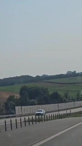 The video shows a highway from a car's perspective. The highway is multi-lane with cars and trucks traveling on it. There are sound barriers along the side of the highway, and behind the barriers are green fields and trees. The sky is clear and blue. The video pans to the right, showing more of the highway and the surrounding landscape.