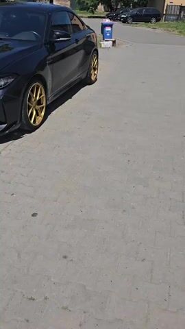 The video shows a black car with gold wheels parked on a paved surface. The car is viewed from the side, with the rear wheel visible. The environment appears to be a street or parking area with some grass and buildings in the background. There is a blue trash bin with a white box next to it near the car. The lighting suggests it is daytime.