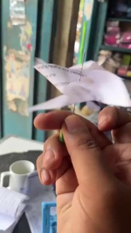 A close-up shot shows a person's hand holding a makeshift pinwheel made from paper and a thumbtack. The pinwheel is spinning rapidly. The background is blurred, but appears to be an indoor setting with shelves stocked with various items.