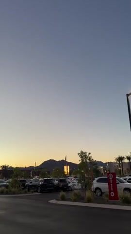 A vertical video captures a parking lot with several cars during dusk. In the distance, mountains are silhouetted against a pale yellow and blue sky. A building with lights on is visible behind the mountains, and a flag flies above it. Palm trees and smaller trees are scattered throughout the parking lot, some with lush green foliage and others with sparser leaves. A red sign with white lettering that says "DRIVE UP" is placed near the right side of the frame. The parking lot pavement is dark and wet in some areas, reflecting the fading light. The video is taken from a low angle, looking up towards the sky.