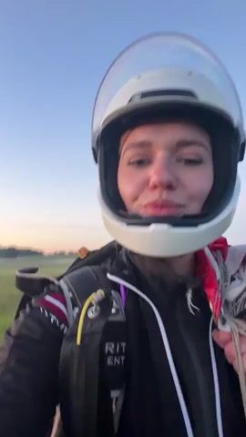 The video features a young woman wearing a white skydiving helmet with a clear visor. She is dressed in a black skydiving suit with red and white accents, and a harness with visible straps and a yellow handle. The woman is smiling and appears excited. She is holding a parachute.

The scene is an open field with green grass under a clear sky. The sun is low on the horizon, suggesting either sunrise or sunset, casting a warm glow on the landscape. There are some trees and possibly buildings visible in the distance. The overall atmosphere is peaceful and serene.

The woman is speaking in Russian, stating that this is her third jump of the day and she's excited. The text on her suit is partially visible and appears to be "SPIRIT SKYDIVING". The time of day is likely early morning or late evening due to the lighting. The location is identified as Limbaži, Latvia. The weather appears to be clear and pleasant.