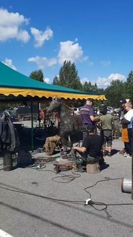 The video shows an outdoor scene, possibly a festival or a public event, with several people working on metal sculptures under a sunny, cloudy sky. In the foreground, a metal horse sculpture is being worked on, with welders and tools around it. To the right, a large geometric metal bear sculpture is visible. People are dressed in casual clothing, some wearing welding gear, including aprons and gloves. Welding equipment, gas cylinders, and metal scraps are scattered around the work area. In the background, there are tents, trees, and other people observing or participating in the event. The overall atmosphere appears to be one of creative construction or artistic display.