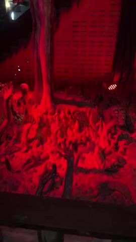 The video shows a dimly lit, outdoor scene, possibly a Halloween decoration or an art installation. The dominant color is a bright, fiery red, cast by red lights that illuminate the entire area. The scene appears to depict a chaotic and disturbing landscape, possibly a representation of hell or a nightmare. Scattered throughout the scene are skeletal figures and grotesque faces, emerging from what looks like molten or burning ground. There are also large, disembodied hands with dark fingernails reaching out from the scene. The overall impression is one of horror and unease, with the red lighting enhancing the sinister atmosphere.