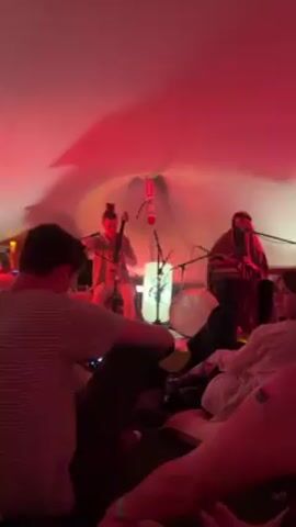 The video shows a musical performance taking place indoors, under a large, conical white tent illuminated with red light. Two musicians are at the front: on the left, a woman with her hair in a bun plays a cello, and on the right, a person with long dark hair, wearing a headscarf and cloak, plays a stringed instrument.  In front of them is a white drum and a decorative tree-like object. Several people are sitting on the floor on rugs and pillows, watching the performance. Some are facing the musicians, while others are turned to the side. The camera focuses on the backs of heads and shoulders of people in the audience. One person has a sticker on their arm. The atmosphere is intimate and informal.