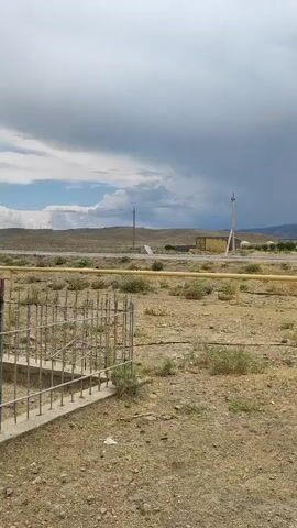 The video shows an outdoor scene with a dirt road and sparse vegetation. In the foreground, there is a section of rebar and concrete construction, partially obscured. A yellow pipe runs horizontally across the middle ground. Further back, several buildings are visible, along with utility poles and a truck. The sky is cloudy, with dark storm clouds gathering in the distance. In the final seconds, a silver SUV is prominently displayed in the foreground on the right side of the frame. The scene appears to be a rural or undeveloped area.