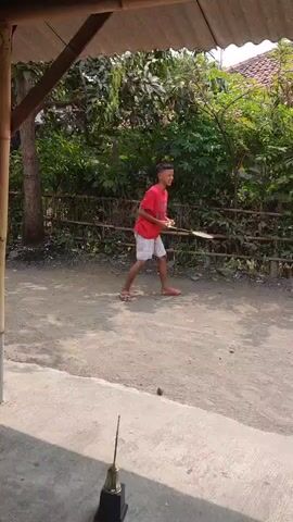 A young person wearing a red t-shirt and white shorts plays badminton outdoors. They hold a racket in their right hand and repeatedly hit a shuttlecock into the air, demonstrating their skill. The setting appears to be a rural area with a dirt path, a fence, and lush green vegetation in the background. A part of a building's roof is visible in the foreground, suggesting the scene is captured from a sheltered spot. The video captures the individual's focus and coordination as they engage in the sport.