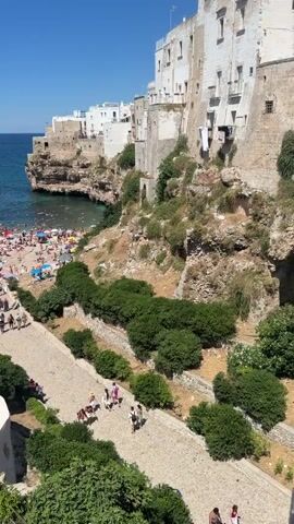 The video shows a crowded beach on a sunny day. People are sunbathing on the sand, swimming in the ocean, and relaxing under umbrellas. Buildings are visible on the cliffs overlooking the beach, and lush green vegetation covers the rocky terrain. The water is a clear blue, and the sky is a vibrant blue with no clouds. The scene depicts a lively and popular tourist destination, with many people enjoying the summer weather and the beautiful coastal scenery.