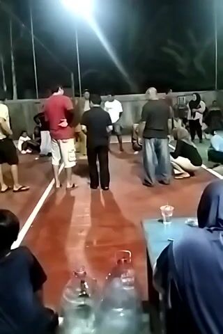 A group of people are gathered on a reddish-brown court, likely outdoors, as evidenced by the palm trees visible in the background and the overhead lighting fixtures. The lighting suggests it is nighttime or dusk. Several individuals are standing, while others are seated or kneeling. In the foreground, two large plastic water bottles are prominently displayed on a table. One person in the foreground appears to be wearing a blue hijab. The standing individuals are casually dressed. One person in the center is wearing a black shirt and pants, and another to their left is in a red shirt and khaki shorts. There's a sense of people watching an event or gathering.
