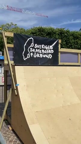 The video shows a skate ramp set up outdoors in an urban environment. A banner at the top of the ramp reads "SVERIGES SKATEBOARD FÖRBUND" with a silhouette of Sweden. The ramp is made of light-colored wood with blue accents. In the background, trees, buildings, and a clear blue sky with some clouds are visible. A construction crane is also visible in the distance. The presence of the skate ramp and banner suggests a skateboarding event or competition is taking place. There are some people visible in the background, but they are not the focus of the video. The scene appears to be set during the daytime, with bright sunlight, indicating fair weather. The overall atmosphere seems to be lively and geared towards an outdoor sporting event.
