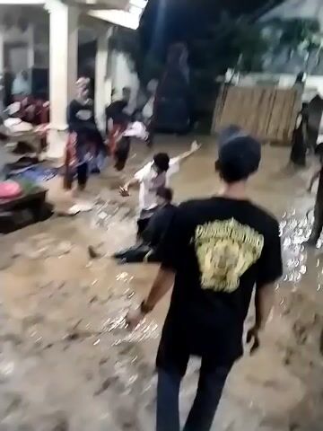 The video depicts an outdoor cultural performance taking place at night in Jenggrik, Indonesia. A group of people, mostly young men, are engaged in a dynamic performance on very muddy ground, which appears to be wet from recent rain. Several participants are actively dancing and moving vigorously, while some are sitting or rolling in the mud as part of the act. Other individuals are gathered around, observing the performance. In the background, illuminated by artificial lights, are houses and what appears to be a bamboo fence. One person in the foreground wears a black t-shirt featuring a golden emblem on the back that reads "PAGUYUBAN SENI REG" with further text resembling "REOG," indicating a traditional Indonesian performing art, likely Reog Ponorogo. The overall scene is lively and engaging.