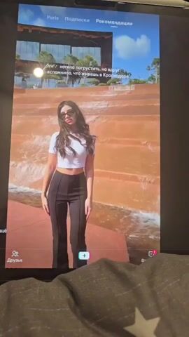 The video begins by showing a tablet screen displaying a social media app. The screen shows a profile page with a user's profile picture on the left and the username "Paris". Below the username are tabs for "Subscriptions" and "Recommendations". The main content of the screen is a video playing, showing a young woman standing on a terrace with a waterfall in the background. She is wearing a white crop top and black pants. The video then transitions to a close-up shot of a red textured wall with visible water streaks. The bottom of the screen shows icons for "Friends" and a plus sign.