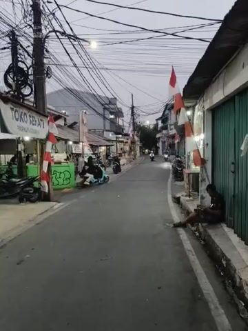 A narrow, paved street is lined with buildings and stalls on both sides. Overhead, a dense network of electrical wires crisscrosses the sky. Several motorbikes are present, with some being ridden down the street and others parked along the sides. A few people are visible, including a man sitting on a concrete ledge on the right side of the street, looking at his phone. On the left side, a sign for "TOKO SEPATI" (Shoe Store) is visible above a stall. Indonesian flags, red and white, are displayed on poles along the street. The lighting suggests it is either early morning or late evening.
