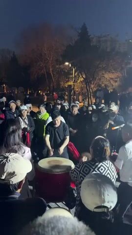 In an outdoor, dimly lit setting, a group of predominantly older adults has gathered around a central figure playing a large drum. The drummer, dressed in a black polo shirt, is actively striking the drum with both hands. The onlookers, many wearing hats, are watching and listening. Some individuals are wearing masks, while others are looking at the drummer with interest. The environment includes trees, streetlights, and a paved area, suggesting a park or public space. The overall atmosphere is one of community engagement and musical performance.