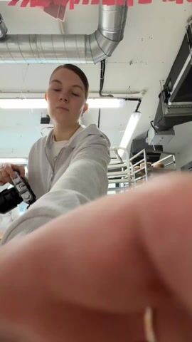 A woman in a gray hoodie and black pants stands at a wooden table, holding a black camera to her eye, apparently taking pictures. On the table are pieces of lace, paper, and various decorative items. Behind her, a metal rack is filled with what appear to be cakes or pastries. In the background, another person wearing a white chef's coat is visible. The room is brightly lit, with metal ductwork and fluorescent lights on the ceiling. Red and pink paper decorations are hung above the table.